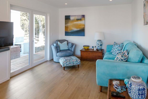 Bright, compact living room with white walls, warm timber flooring, blue and turquoise upholstered seating, patterned cushions, a wooden sideboard, and abstract artwork. Full-height glazed French doors open to a timber deck, bringing in abundant natural light.
