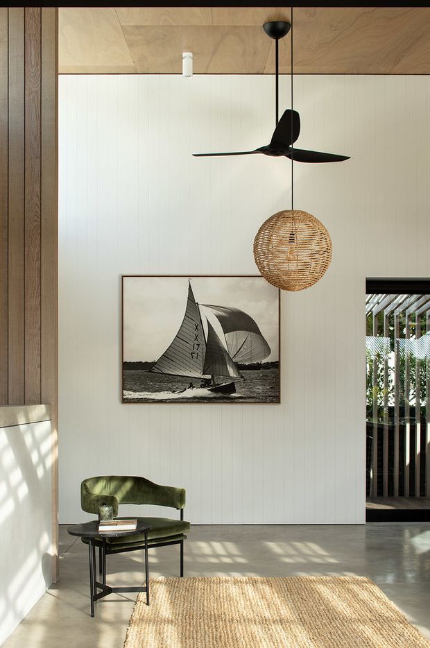 A bright, double-height interior corner with vertical timber wall framing, a warm timber-paneled ceiling, white vertical wall cladding, and a polished concrete floor. A black sculptural ceiling fan and woven pendant light hang above a vintage green lounge chair and small black side table. A framed black-and-white sailing photograph provides a focal point, while a woven natural-fibre rug and strong window shadows add texture and warmth. An opening at right reveals a covered outdoor walkway or glazed terrace.