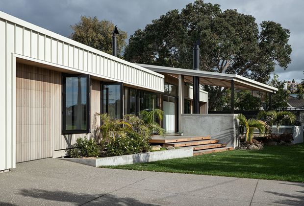 A low, single-storey contemporary house viewed from the front garden and paved driveway. The facade combines pale vertical metal cladding with timber-lined recessed walls, extensive dark-framed glazing, a broad shallow roof overhang, and a raised timber deck with concrete planter and privacy wall. Tropical planting and mature trees surround the house.
