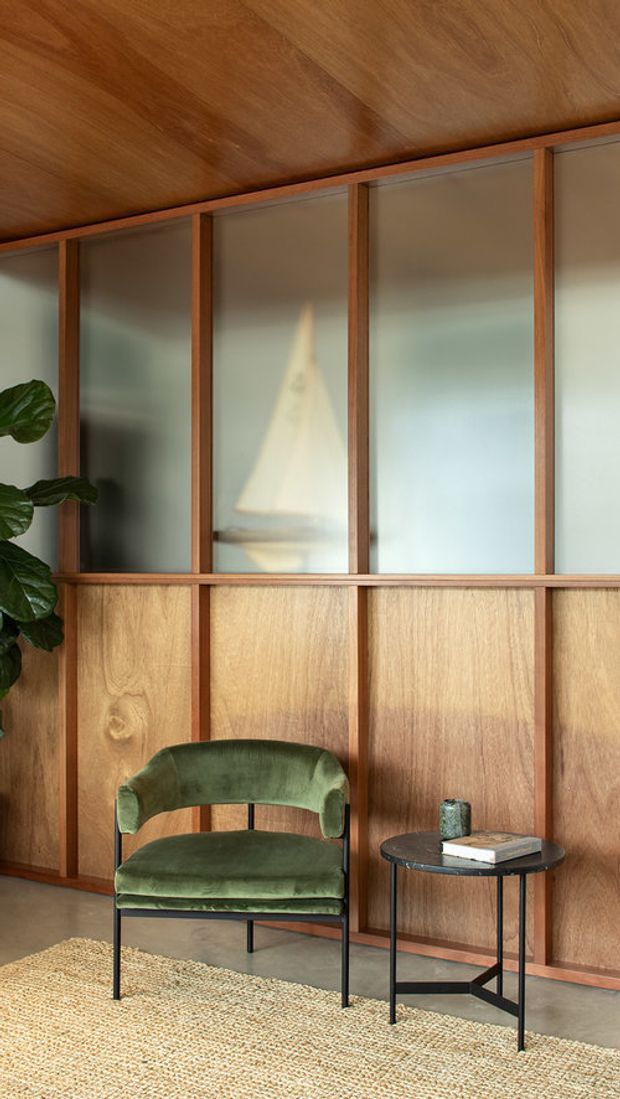 A warmly lit interior vignette featuring a green upholstered lounge chair and small black side table in front of a full-height timber and frosted-glass partition wall. The wall has rich timber framing, lower timber panelling, and translucent upper panels. A woven natural-fibre rug, leafy plant, and subtle decorative objects add texture and a calm mid-century character.