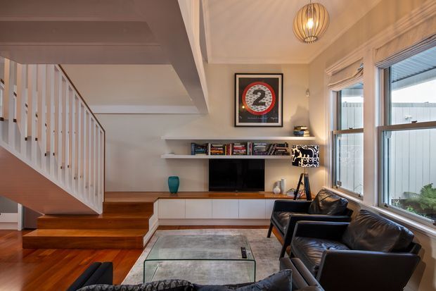 A warm, compact contemporary living room with a timber staircase, built-in white entertainment cabinetry and shelving, wall-mounted television, black leather armchairs, a glass coffee table, and a large area rug. Tall windows with Roman blinds bring in daylight, while a pendant light and table lamp add warmth. A framed graphic artwork hangs above the media unit.