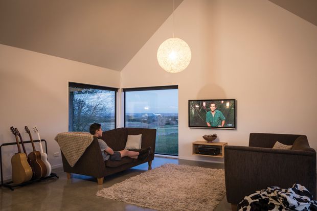 A warm, compact living room with a vaulted ceiling, large corner windows overlooking a distant landscape, a central spherical pendant light, two dark upholstered sofas, a shag rug, a wall-mounted television, and a guitar stand. The room has a relaxed contemporary feel with polished concrete-style flooring and neutral finishes.