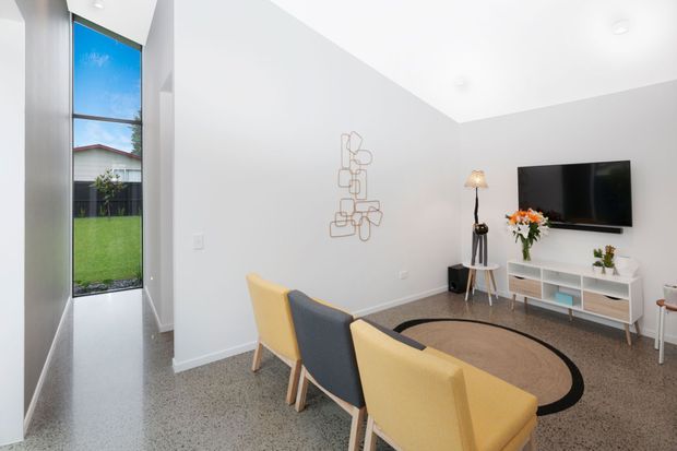 Bright contemporary living room with a high sloping ceiling, polished concrete or terrazzo-like flooring, white walls, and tall vertical glazing opening to a small lawn. A wall-mounted television sits above a low white media console, with colourful upholstered lounge chairs arranged around a circular rug and a small side table. Minimal metal wall art and simple decorative styling complete the space.
