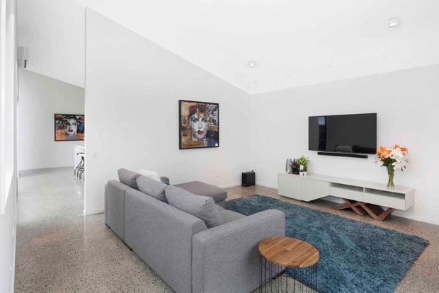 Bright contemporary living room with white walls and ceiling, polished grey-speckled flooring, a large grey sectional sofa, blue area rug, white low-profile media console, wall-mounted television, timber round side table, artwork and decorative flowers. An open hallway or adjacent space is visible at left.