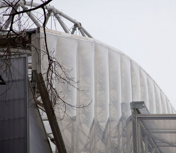Close-up exterior view of a large curved translucent building envelope with visible structural steel framing and tubular roof supports. The facade appears to use translucent polycarbonate or membrane-like panels, with an attached glazed or translucent canopy at lower right. Leafless branches partially obscure the foreground.