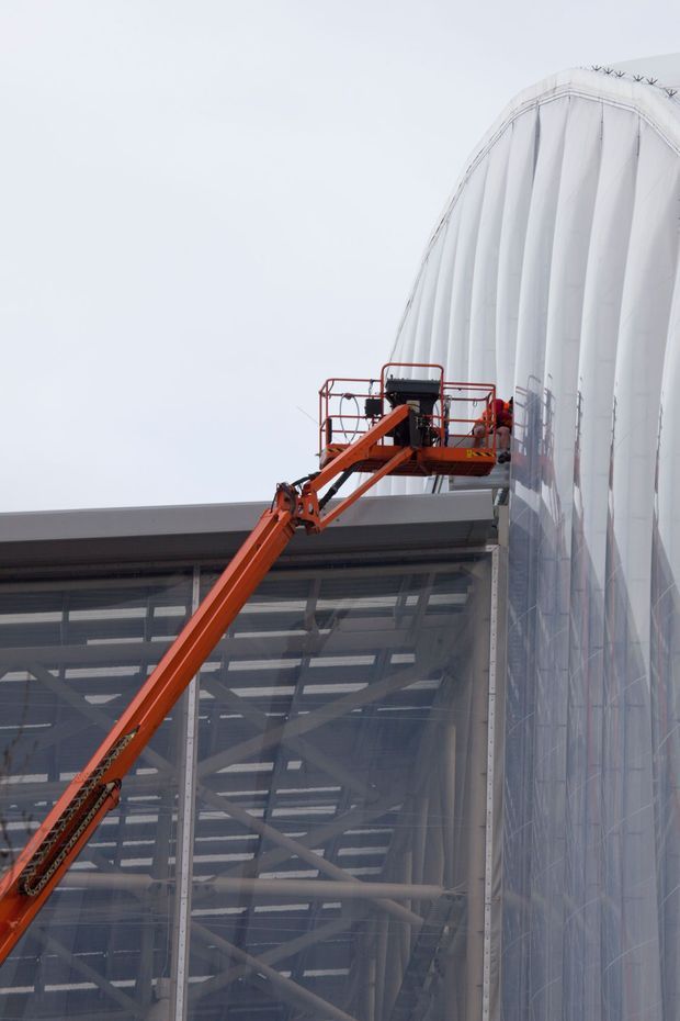 Exterior construction photograph showing a large industrial or commercial building with a curved, translucent white membrane facade and extensive glazed structural framing. An orange articulated boom lift is positioned against the facade, indicating construction or maintenance work. The image is dominated by the building envelope and construction equipment, with an overcast pale sky in the background.