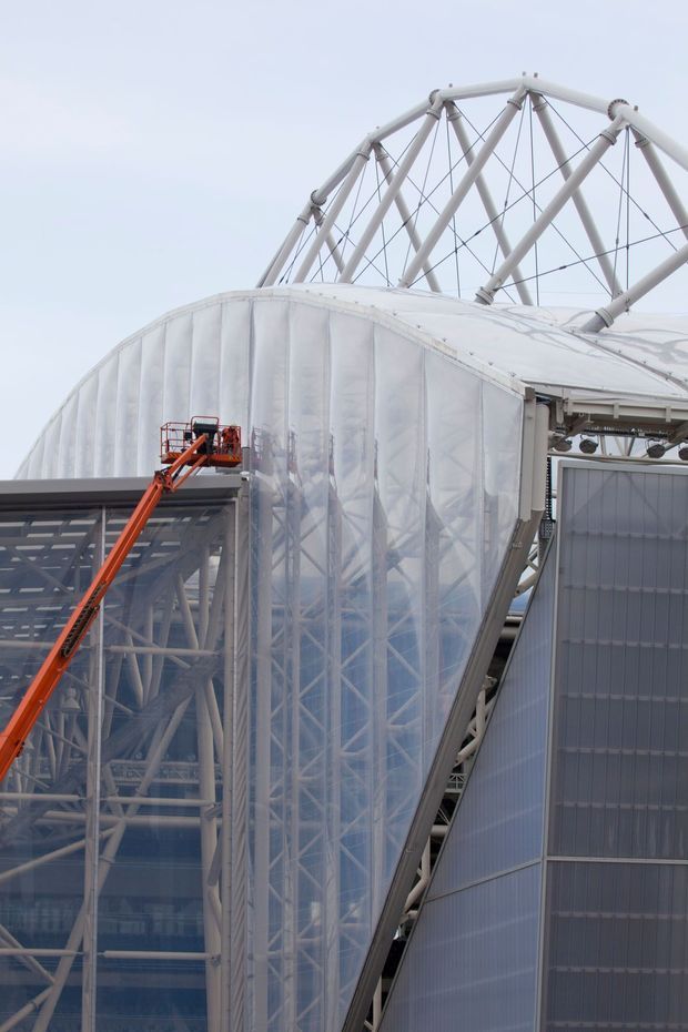 Close-up exterior view of a large contemporary steel-framed building with translucent white membrane or polycarbonate cladding, exposed tubular roof trusses, and extensive glazing. An orange elevated work platform is visible beside the facade, suggesting construction or maintenance. The image emphasizes the building's structural framework and lightweight envelope.