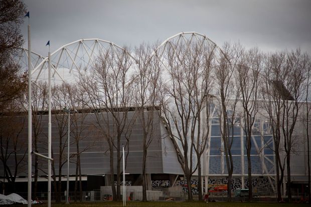 Exterior view of a large contemporary commercial or public building behind a row of leafless trees. The facade combines dark grey cladding, extensive glazed sections, and prominent white arched truss structures rising above the roofline. Flagpoles and paved landscaping are visible in the foreground under an overcast sky.