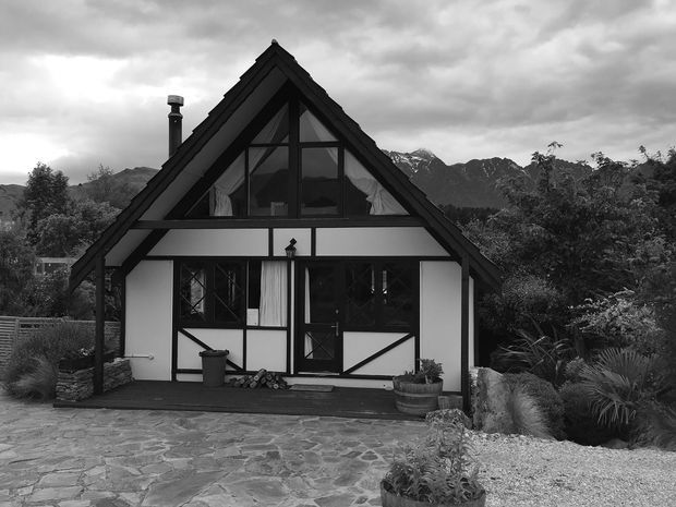 Black-and-white exterior photograph of a compact alpine-style cottage with a steep front-facing gable roof, dark timber framing, white infill walls, and upper-level glazing. The house is set against a mountainous landscape with trees and landscaped planting, with a stone-paved forecourt and small entry porch in front.