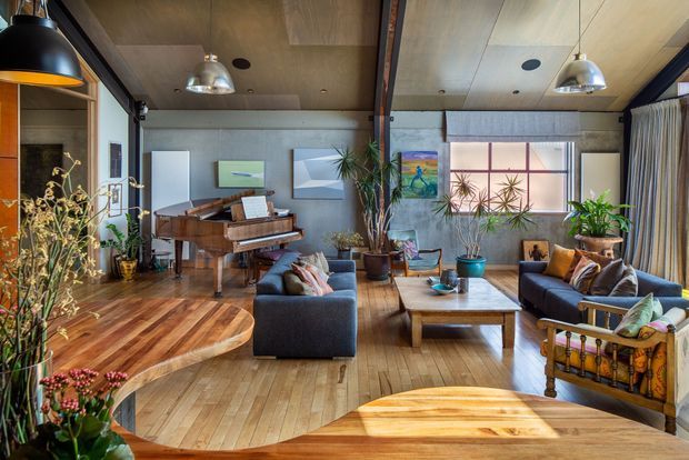 Warm, eclectic open-plan living space with a grand piano, dark upholstered sofas, timber coffee table, abundant indoor plants, artwork, and extensive timber flooring. The room features a distinctive curved raised timber platform around the piano and a timber-lined ceiling with exposed structural beams, pendant lighting, and large glazed openings.