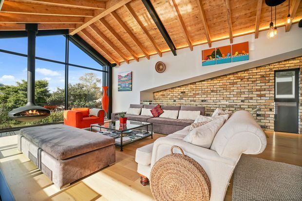 Spacious, warm living room with a dramatic exposed timber pitched ceiling, exposed rafters, a full-height glazed corner, suspended fireplace, exposed brick feature wall, timber flooring, and layered contemporary seating. The room has strong indoor-outdoor views and abundant natural light, with colourful artwork and orange accent chairs adding contrast.
