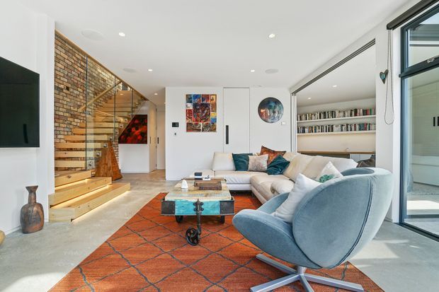 Bright open-plan living room with a white ceiling and walls, exposed brick feature wall, timber stairway with glass balustrade, built-in white shelving, and large black-framed glazing. The room is furnished with a pale sectional sofa, sculptural blue lounge chair, glass-topped coffee table, and a large rust-orange patterned rug. Contemporary artwork and books add colour and character.