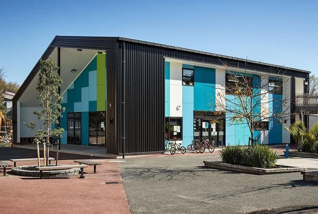 Exterior view of a contemporary multi-storey commercial building with a steeply sloped roof, dark vertical ribbed cladding, and a bold turquoise, blue, white, and lime geometric facade. Large glazed entry and ground-floor openings face a paved landscaped forecourt with benches, planters, and bicycle racks.