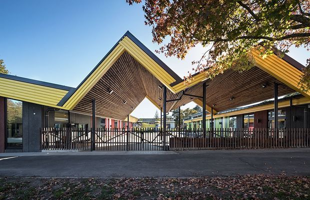 Exterior view of a contemporary low-rise building complex with a dramatic series of intersecting pitched roof forms. The facade combines dark cladding, bright yellow-green horizontal cladding, timber-lined soffits, extensive glazing, and dark metal fencing. A covered entrance or courtyard is prominently framed by the peaked roof, with paved areas in front and mature tree canopy overhead.