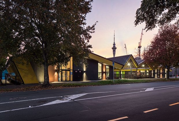 Street-level dusk view of a contemporary low-rise civic or commercial building with bold angular gabled roof forms, dark facade panels, extensive glazing, and bright linear lighting. Mature street trees frame the building, while road markings and construction cranes are visible in the foreground and background.