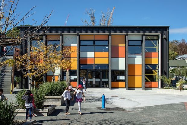 Exterior view of a contemporary multi-storey building with a dark ribbed facade and tall grid-like window walls filled with bold orange, yellow, red, white, and blue panels. The entrance opens onto a paved forecourt with landscaping and people nearby. The composition has a strong graphic, institutional or commercial character.