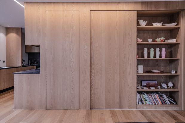 A warm, contemporary open-plan kitchen and built-in storage wall clad in light timber veneer. The central full-height cabinetry conceals storage or appliances, while an open display shelving unit holds ceramics and books. A compact kitchen with dark benchtops is visible to the left, with matching timber flooring and subtle linear ceiling lighting.