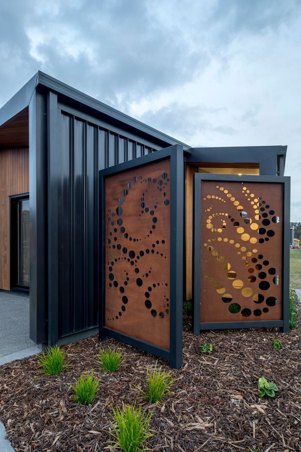 Exterior view of a contemporary single-storey building with dark vertical metal cladding, timber-lined soffits and warm timber wall sections. Two freestanding rust-toned perforated metal privacy screens with circular patterns define the landscaped entrance, set among mulched garden beds and ornamental grasses under an overcast sky.