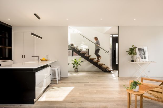 Bright open-plan contemporary interior showing a kitchen, living area, and sculptural open-riser staircase. The kitchen has white cabinetry, a dark island, and a pale timber-look floor. A glazed stair balustrade, indoor plants, and light neutral finishes create an airy, minimalist setting.