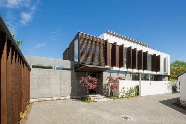 Exterior view of a contemporary multi-level residence from a paved driveway or courtyard. The home features crisp white rendered volumes, exposed concrete wall panels, dark timber vertical screens and horizontal timber sun-shading, extensive glazing, and a projecting upper-level box. Minimal landscaping and sculpted Japanese maple trees frame the entry.