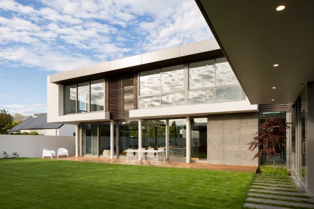 A contemporary two-storey residential house viewed from a landscaped lawn and covered outdoor area. The building has a clean rectilinear form, extensive floor-to-ceiling glazing, white rendered or panelled surfaces, dark horizontal sun-shading, and a large grey stone-clad wall. A paved path crosses the lawn toward the covered patio, with outdoor dining furniture visible beneath the upper level. The image has a bright, airy architectural photography style.