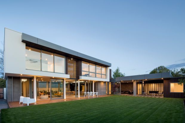 Exterior view of a large contemporary two-storey residence with a clean rectilinear form, extensive floor-to-ceiling glazing, dark horizontal roof and fascia elements, timber screening, and a connected single-storey wing. The home opens onto a broad manicured lawn with timber decking and outdoor seating, captured in soft evening light.