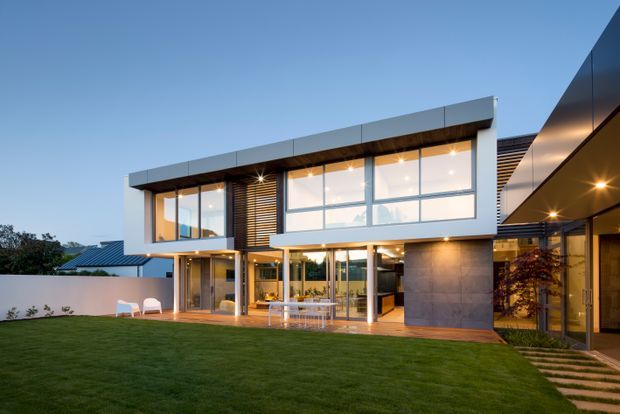 Exterior view of a contemporary two-storey residence at dusk, with a flat-roofed main volume, extensive floor-to-ceiling glazing, aluminium-framed windows, timber-toned horizontal screening, and a covered outdoor entertaining area opening onto a manicured lawn. A connected side wing and illuminated entry walkway are also visible.