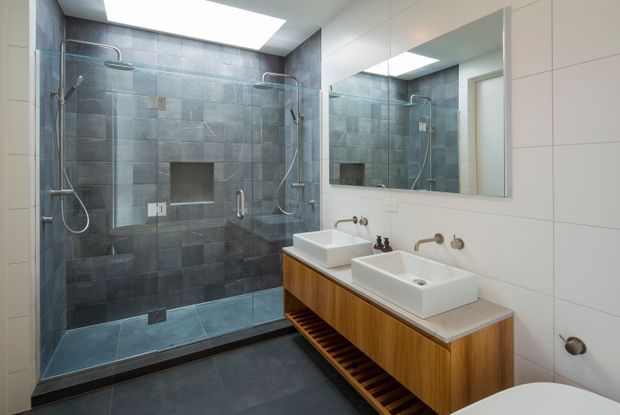 Contemporary bathroom with a large frameless glass double shower, dual white vessel basins on a timber vanity, expansive wall mirror, dark grey tiled surfaces, and a bright skylight.