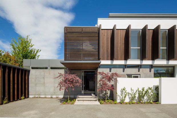 Contemporary multi-storey residential facade viewed from the street, with clean white rendered walls, dark timber vertical fins, a timber louvre screen over the entry, extensive glazing, and a concrete boundary wall. A paved driveway and landscaped entry planting complete the modern composition under a bright blue sky.