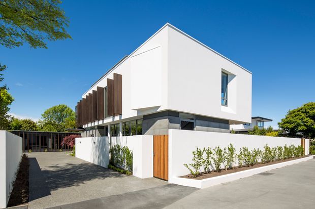 A contemporary two-storey residential house viewed from the street, with crisp white rendered walls, a steep gabled roof form, narrow vertical windows, a dark glazed lower level, and a screened balcony or upper-level opening lined with vertical timber elements. A white boundary wall, timber pedestrian gate, driveway, and landscaped planting define the frontage.