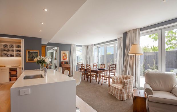 Open-plan contemporary interior combining a white kitchen with a large island, dining area, and bright living space. The room features extensive floor-to-ceiling glazing with curtains, neutral carpeting, dark grey and white walls, timber dining furniture, upholstered armchairs, recessed ceiling lights, and a display nook with built-in shelving.