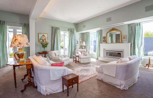 Bright, spacious formal living room with pale sage-green walls, tall windows dressed in soft green curtains, white slipcovered sofas and armchairs, a central patterned rug, traditional timber side tables, decorative artwork, and a classic white fireplace with an ornate overmantel mirror. The room has abundant natural light and refined traditional detailing.