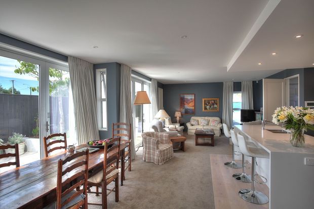 Open-plan living, dining, and kitchen interior with abundant floor-to-ceiling glazing, full-height curtains, recessed ceiling lighting, and a neutral carpeted floor. The dining area has a timber table and ladder-back chairs, while the living area contains upholstered seating and a coffee table. A large light-coloured kitchen island with bar stools occupies the right side, creating a connected, spacious family living zone.
