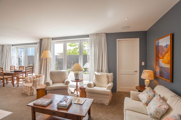 A bright, traditionally styled open-plan living and dining room with large windows, floor-length curtains, upholstered armchairs and sofa, a wooden coffee table, table lamps, framed artwork, and a dark blue-grey feature wall. The dining area is visible at the left, while the main focus is the comfortable living space.