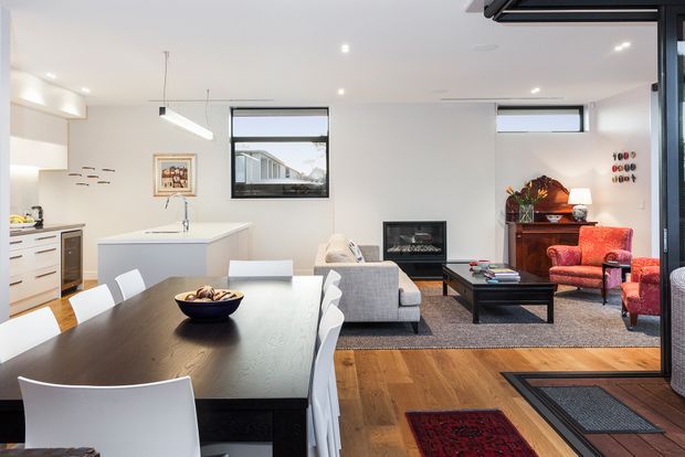 Open-plan contemporary living, dining and kitchen area with a dark timber dining table, white dining chairs, compact white kitchen island and cabinetry, grey sofa, black coffee table, fireplace, and a red upholstered armchair. Large windows and glazed doors bring in daylight, while timber flooring and layered rugs add warmth.