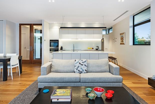Bright open-plan contemporary living area with a grey upholstered sofa in the foreground, a black coffee table, and a white minimalist kitchen with island behind it. Warm timber flooring, white cabinetry, integrated appliances, black-framed windows, recessed lighting, and a dining area create a clean, modern family interior.