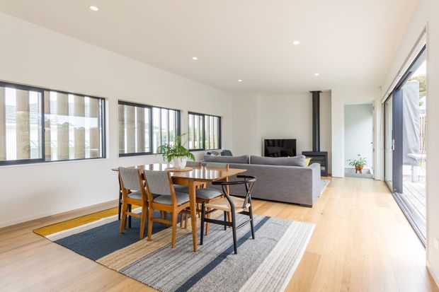 A bright, open-plan contemporary living and dining area with pale timber flooring, white walls, large black-framed windows, and extensive sliding glass doors. A timber dining table with mixed chairs sits on a large striped rug, while a grey sofa faces a compact black fireplace and hearth. The room opens toward an outdoor area through full-height glazing.