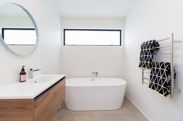 Bright contemporary bathroom with a freestanding white bathtub beneath a narrow horizontal window, a wall-mounted timber vanity with white benchtop, round mirror, and stainless-steel heated towel rail holding patterned towels. The room has light textured walls, pale tiled flooring, and black-framed glazing.