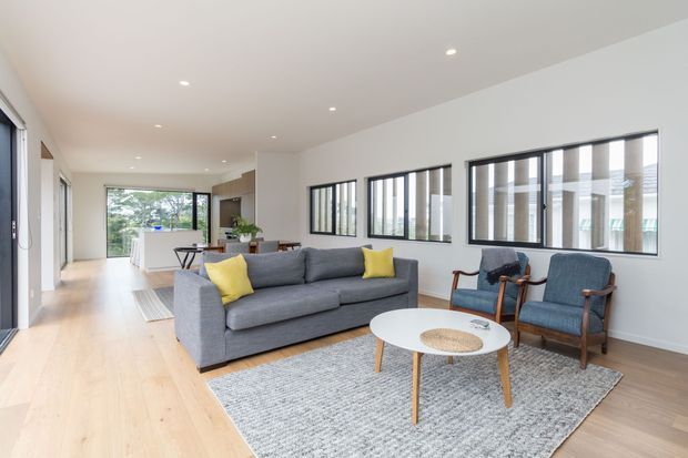 Bright open-plan living room with a grey sofa, yellow accent cushions, two blue upholstered armchairs, a round white coffee table, and a large grey rug. Pale timber flooring, white walls and ceiling, recessed downlights, and extensive black-framed interior glazing create a clean contemporary feel. A kitchen and dining area are visible in the background.