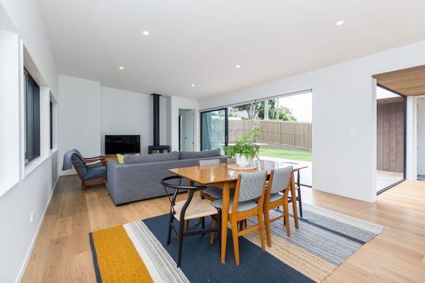 Bright open-plan interior combining a dining area and living room. The space features pale timber flooring, white walls and ceiling, recessed downlights, large windows and glazed openings, a grey sofa, a timber dining table with mixed chairs, and layered striped rugs. A partially visible timber-lined outdoor or semi-enclosed area is seen through the opening on the right.