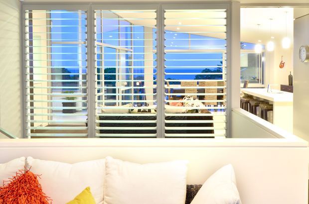 Bright contemporary interior viewed from behind a white sofa, looking through wide louvred windows toward an outdoor terrace and blue sky. A white kitchen or dining area with pendant lighting is visible to the right, creating an open-plan residential setting. The space uses a predominantly white palette with strong natural light and coastal-style louvres.