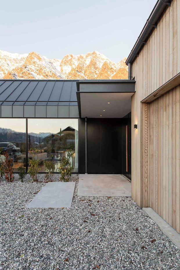 Contemporary mountain-home exterior showing a sheltered entry courtyard with dark metal roofing and fascia, warm vertical timber cladding, large glazed windows, concrete stepping slabs, and a gravel landscape. Snow-covered alpine mountains are visible beyond the house.