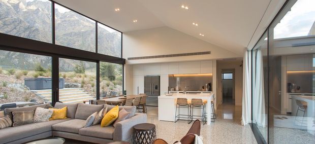 A bright open-plan contemporary interior combining a living room and kitchen, with a large sectional sofa in the foreground, a white island with bar stools, and extensive floor-to-ceiling glazing. Dark-framed windows overlook a dramatic mountain landscape and landscaped outdoor steps. The room has a high sloping ceiling, recessed lighting, polished light terrazzo-like flooring, and warm understated finishes.