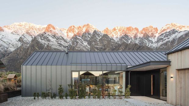 Contemporary single-storey house with a long, low metal-clad roof and dark standing-seam facade, set against dramatic snow-capped alpine mountains. Floor-to-ceiling glazing opens onto a gravelled landscaped forecourt, while a timber-clad wing and recessed dark entry add contrast.
