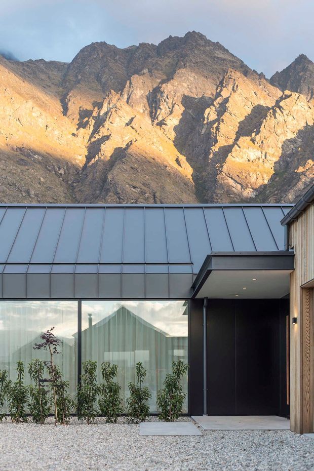 A contemporary mountain-facing building exterior with a steep standing-seam metal roof, extensive glazed wall, dark recessed entry canopy, and vertical timber cladding. A dramatic rugged alpine mountain range dominates the background, while a gravel forecourt and planted glazing edge frame the lower portion of the image.