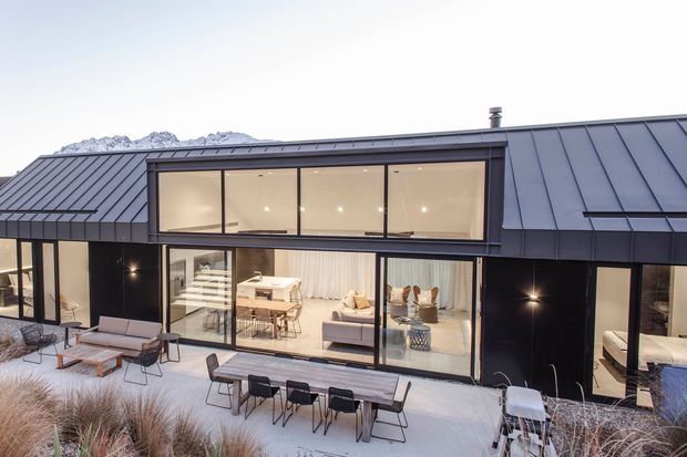 Contemporary single-storey residence with a dark metal pitched roof and black facade, viewed from an outdoor terrace. Large floor-to-ceiling glazed openings reveal a bright open-plan living and dining area, while outdoor dining furniture and ornamental grasses occupy the foreground. Snow-capped mountains are visible in the background.
