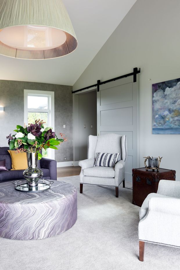 A refined living room featuring soft grey carpeting, a purple sofa, light upholstered armchairs, a sculptural round coffee table with a dramatic wavy-patterned surface, and a vintage dark wood trunk side table. Grey textured walls contain a large sliding barn-style door with black hardware, while a large pleated pendant light and abstract artwork add visual interest.