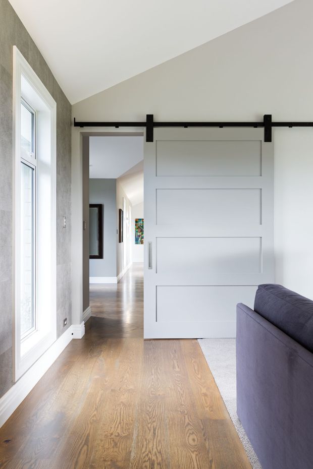Bright contemporary interior showing a large white paneled barn-style sliding door on black hardware, opening from a living area into a hallway. The space has warm timber flooring, pale walls, a tall white-framed window, and the edge of a dark upholstered sofa.