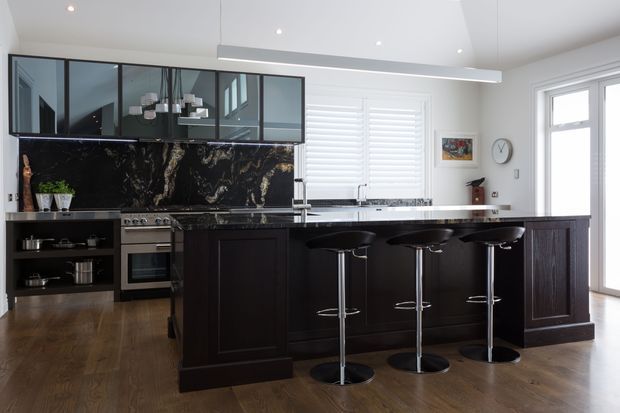 A spacious, refined kitchen with dark timber cabinetry, a large island with seating for four, a dramatic dark marble-look splashback, and integrated stainless-steel appliances. Black-framed glazed upper cabinetry, white walls, plantation shutters, pendant lighting, and timber flooring create a high-contrast contemporary interior.