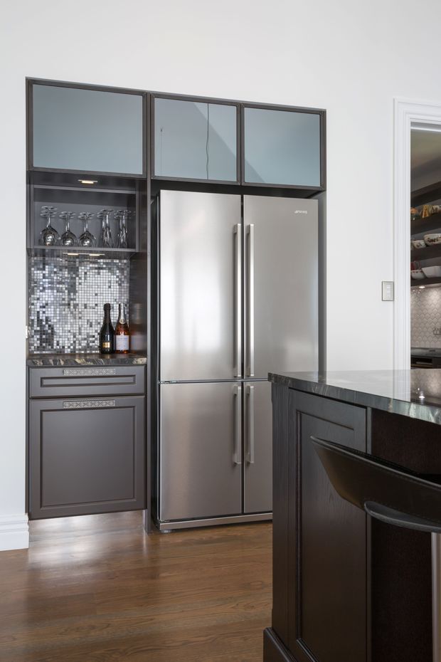 A close-up view of a refined kitchen featuring dark charcoal built-in cabinetry, a large stainless-steel French-door refrigerator, glass-front upper cabinets, and a decorative mosaic tile backsplash with integrated glassware storage. A dark stone countertop and island with seating are visible in the foreground, while a pantry or adjacent kitchen area is partially visible through the doorway.